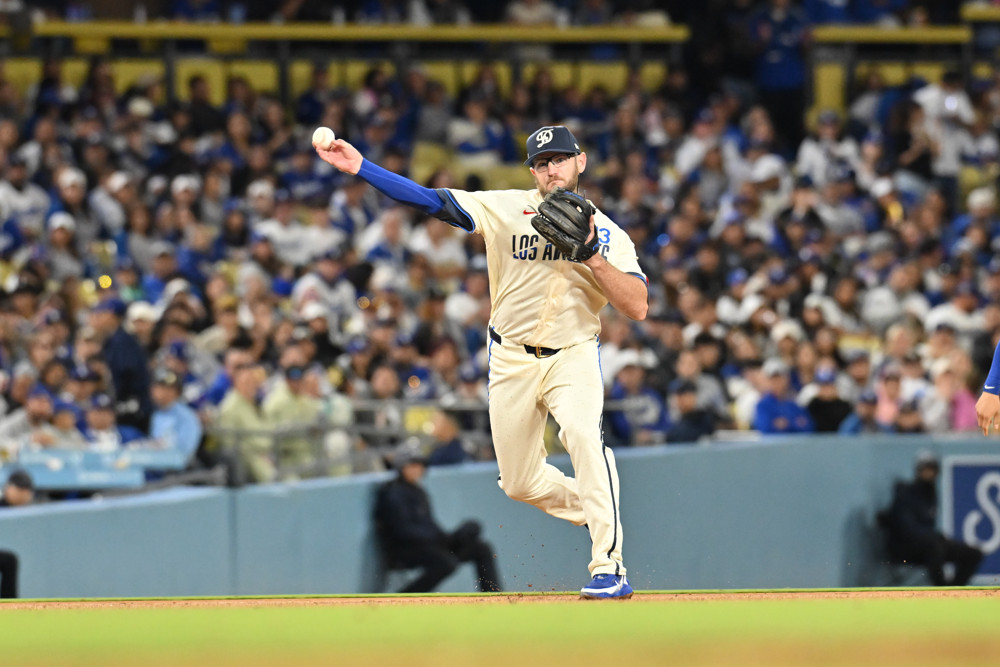 Max Muncy, wearing a white baseball uniform with blue cap and undershirt, prepares a throw to first base from third base.