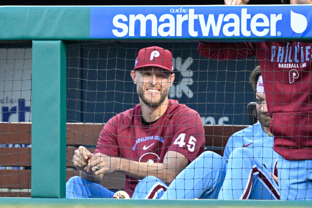 Zack Wheeler, wearing a red Phillies t-shirt and light blue baseball uniform pants, along with a Phillies cap, sits comfortably in the Phillies dugout with a smile on his face.