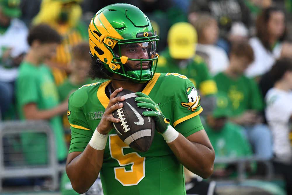 Dante Moore, in green and yellow jersey and green and yellow helmet, drops back to pass, holding the football in front of him.